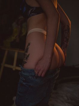 A sensual and artistic side shot of a tattooed woman adjusting denim jeans indoors.