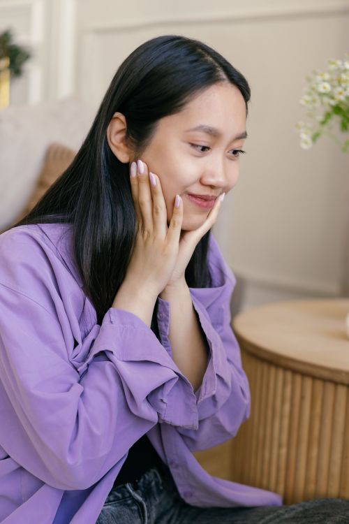 Free Asian woman smiling gently with hands on face, sitting indoors with soft lighting. Stock Photo