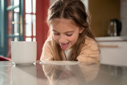 Little Girl Sticking out Tongue Leaning towards Yogurt Spilled on Table