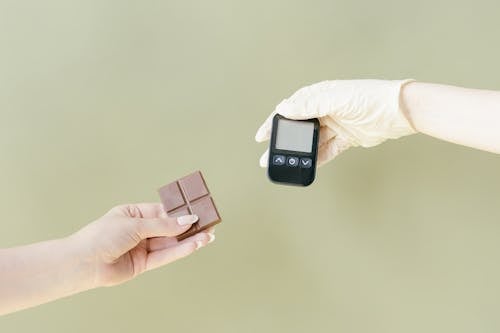 Close-up of One Hand Holding Chocolate and the Other Wearing a Glove and Holding a Glucometer 