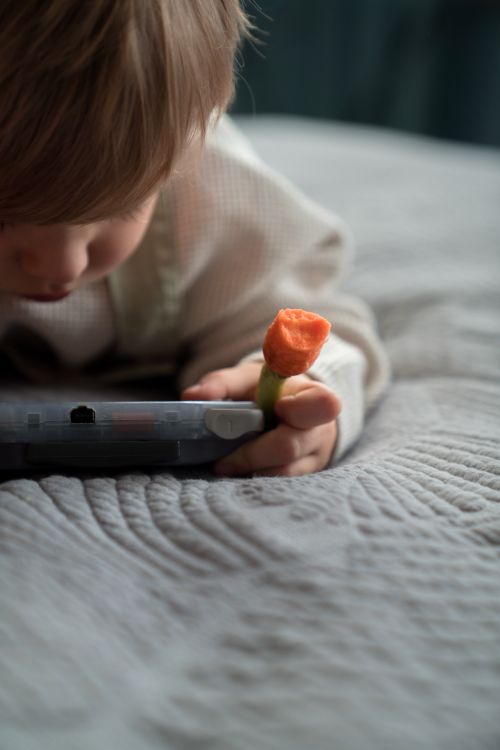 Free A child enjoys handheld gaming while holding a carrot stick indoors. Stock Photo