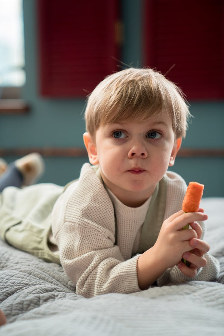 A Boy Eating A Carrot On The Bed