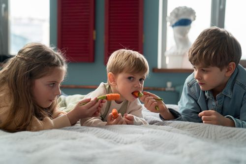 Free Kids lying on bed sharing fresh carrots in a cozy indoor setting. Stock Photo