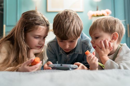 Free Three kids enjoying video games and snacking on carrots indoors, showcasing childhood togetherness. Stock Photo