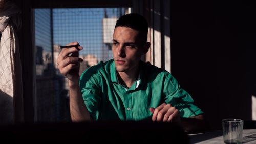 Free A man in a green shirt thoughtfully holding a pencil by a sunlit window. Stock Photo