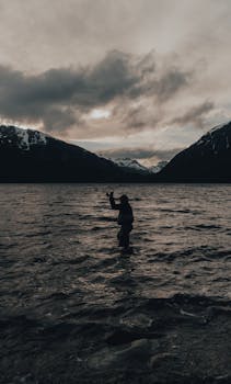 A lone fisherman casts his line in a serene mountain lake, silhouetted against a moody sky.