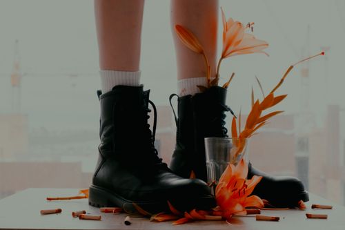 Free Close-up of black boots with vibrant orange lilies and scattered cigarette butts indoors. Stock Photo