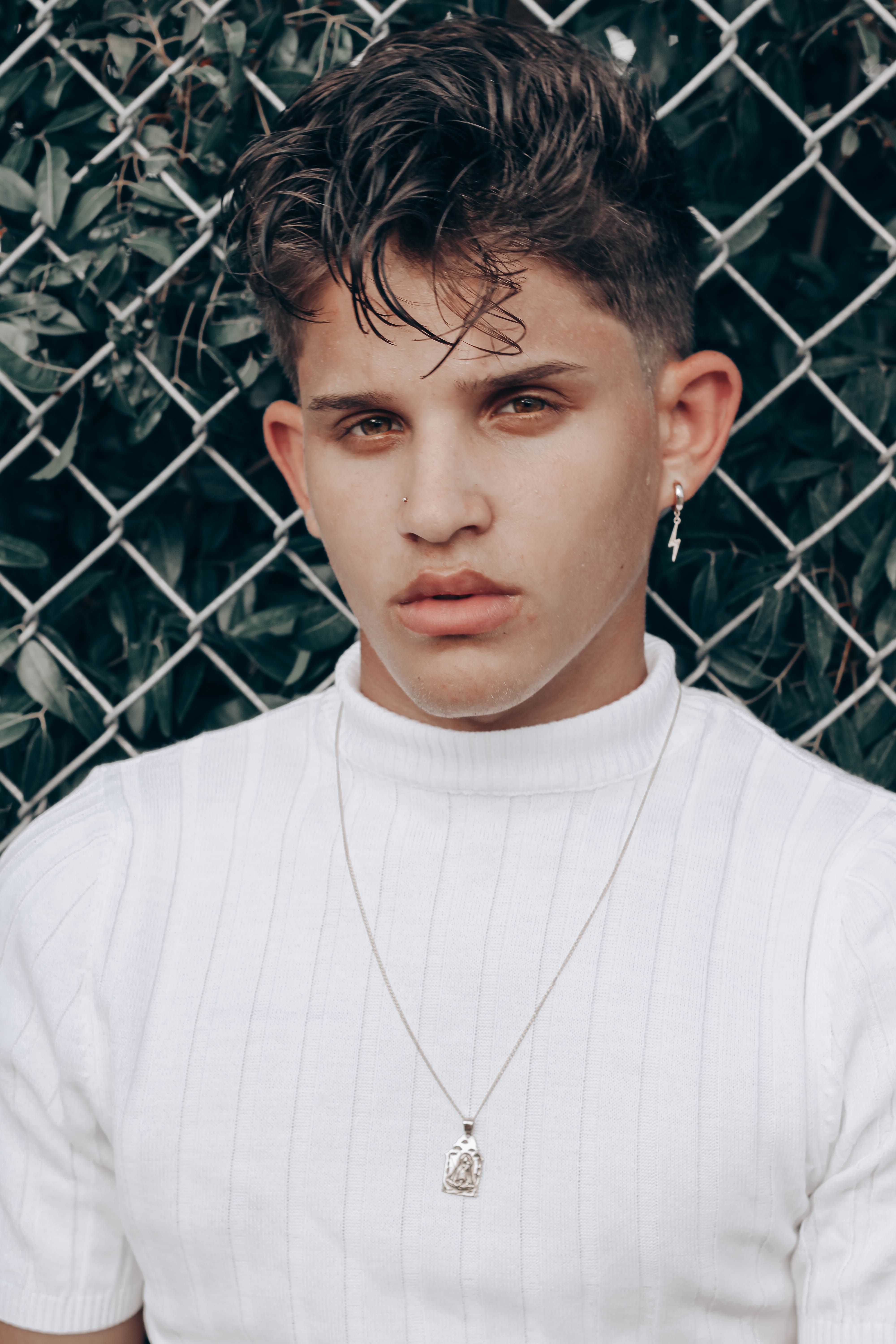Portrait of a fashionable young man with earrings and necklace against a chain-link fence.