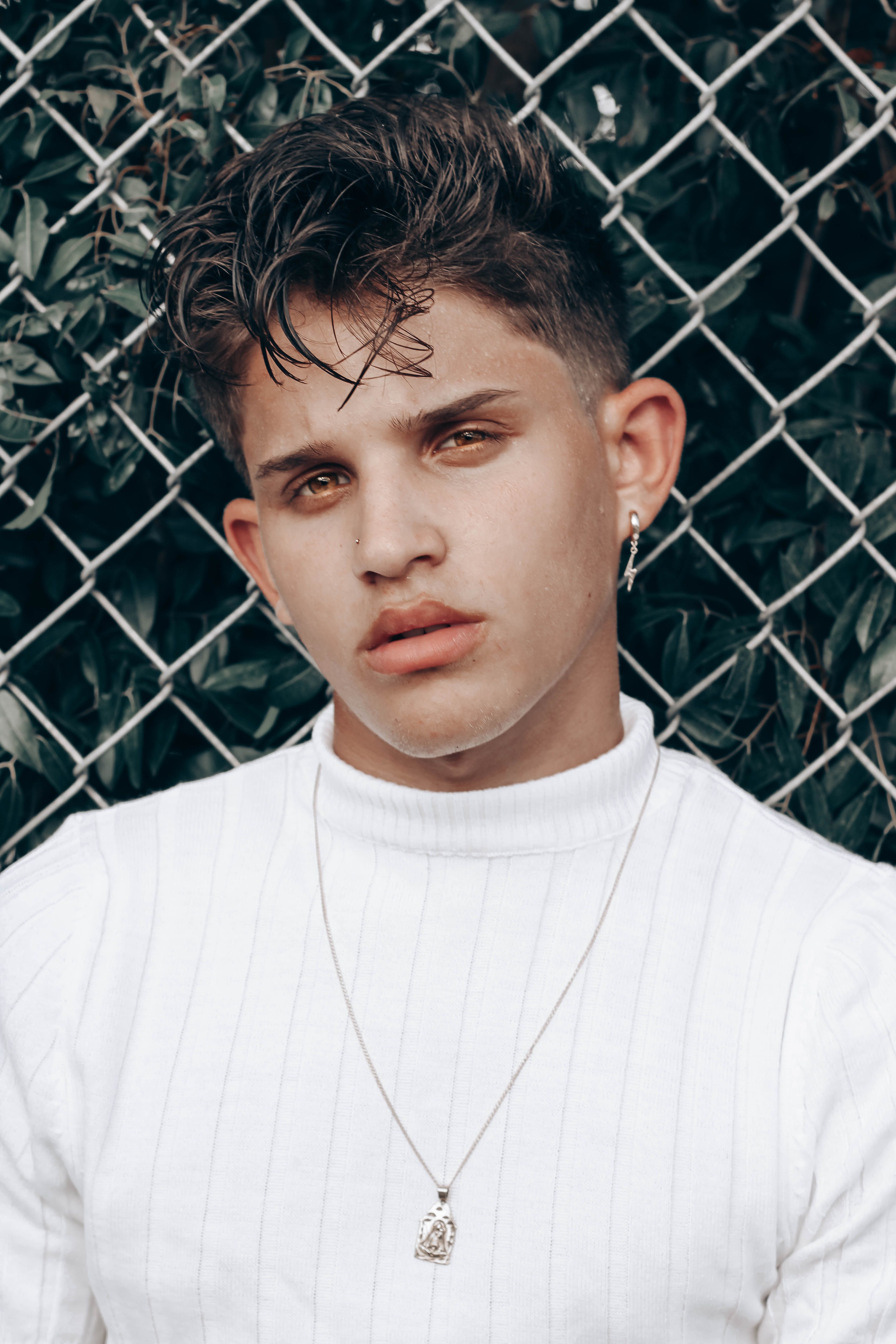 Portrait of a young man with a trendy hairstyle and earrings in front of a chain-link fence.