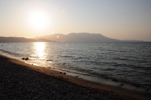 Serene morning scene of a rocky shoreline and gentle waves at sunrise, with mountain silhouettes.