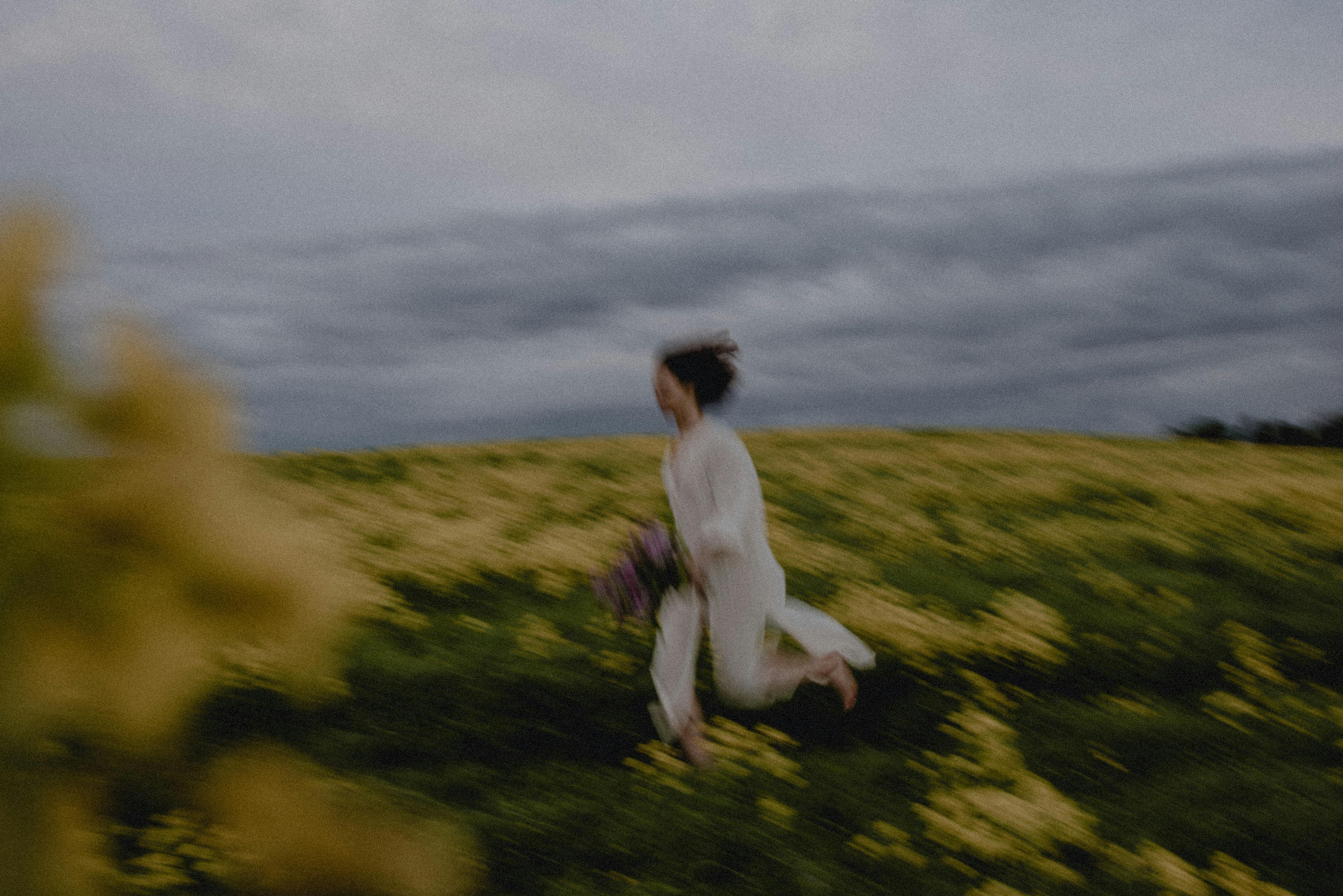 Artistic blur of a woman running, capturing movement amidst a field of yellow wildflowers under cloudy skies.