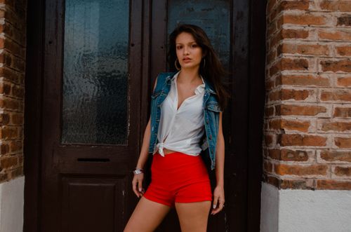 Free Stylish woman in denim vest and red shorts poses by a wooden door, exuding modern fashion vibes. Stock Photo