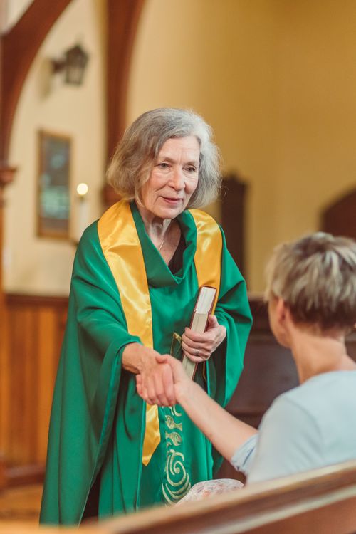 Free An elderly female clergyperson in green robes shakes hands with a visitor inside a church. Stock Photo