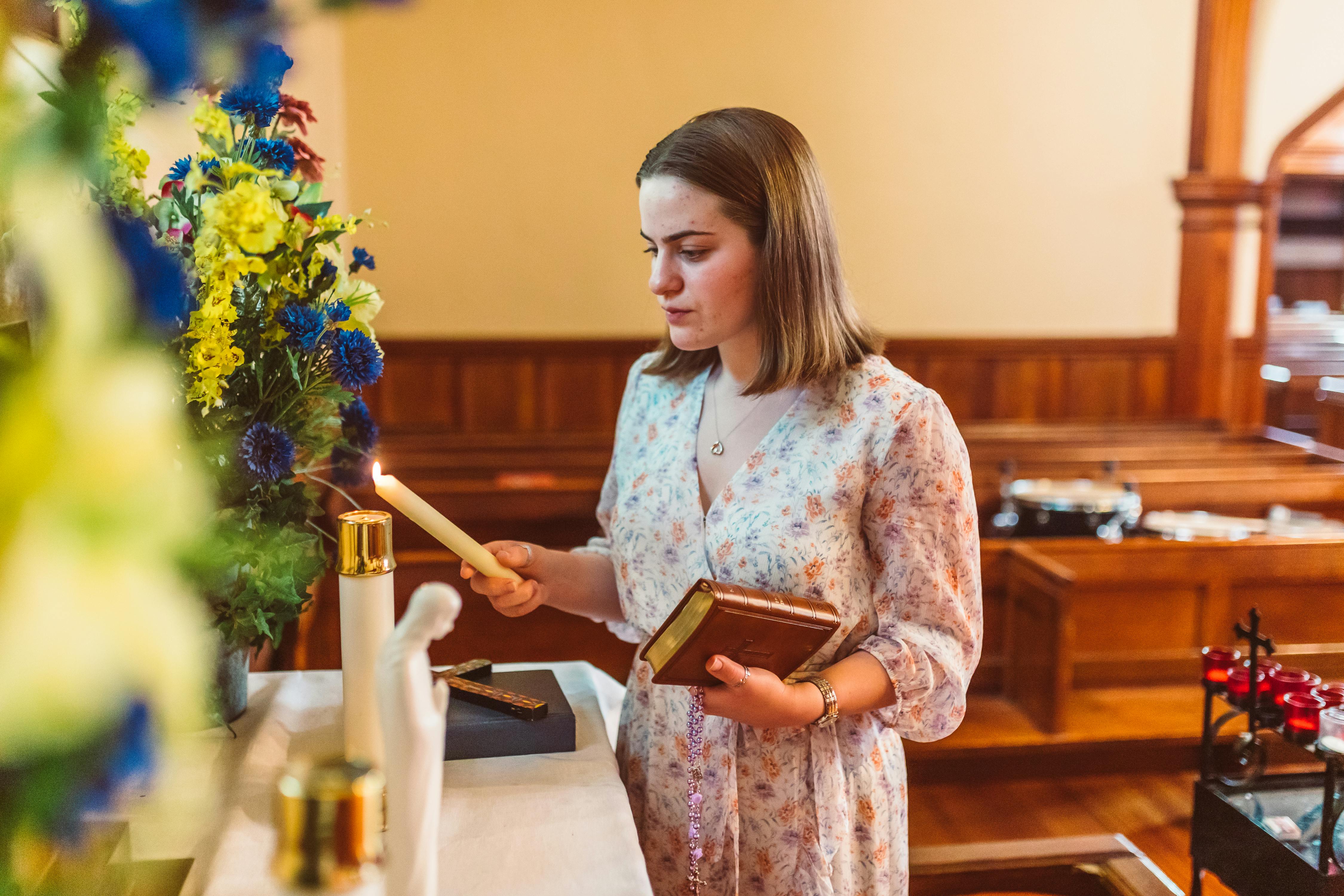 Young caucasian woman lights a candle in a church holding a book, floral dress.