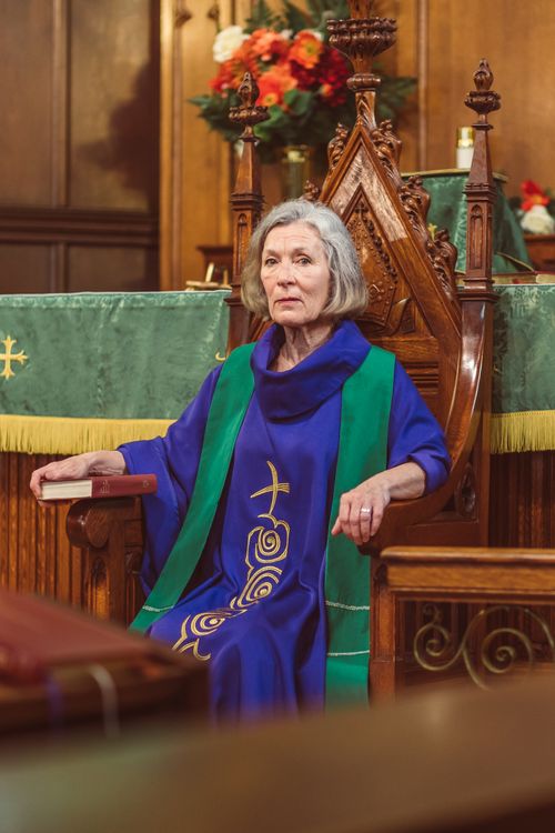 Free Elderly female clergy sitting in ornate chair inside a church, wearing a cassock. Stock Photo