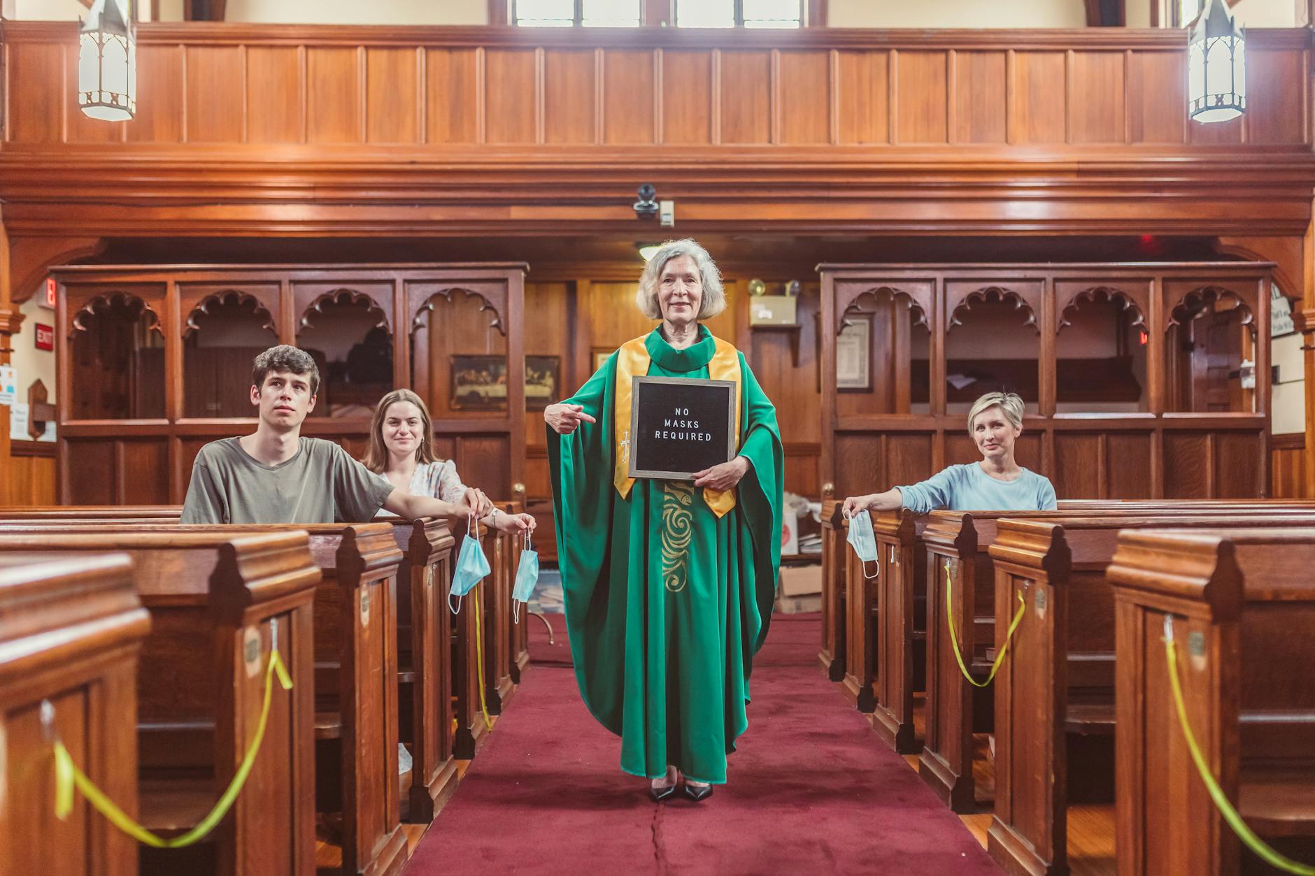 A group in a church pew with a woman holding a 'No Masks Required' sign, indicating a change in safety measures.