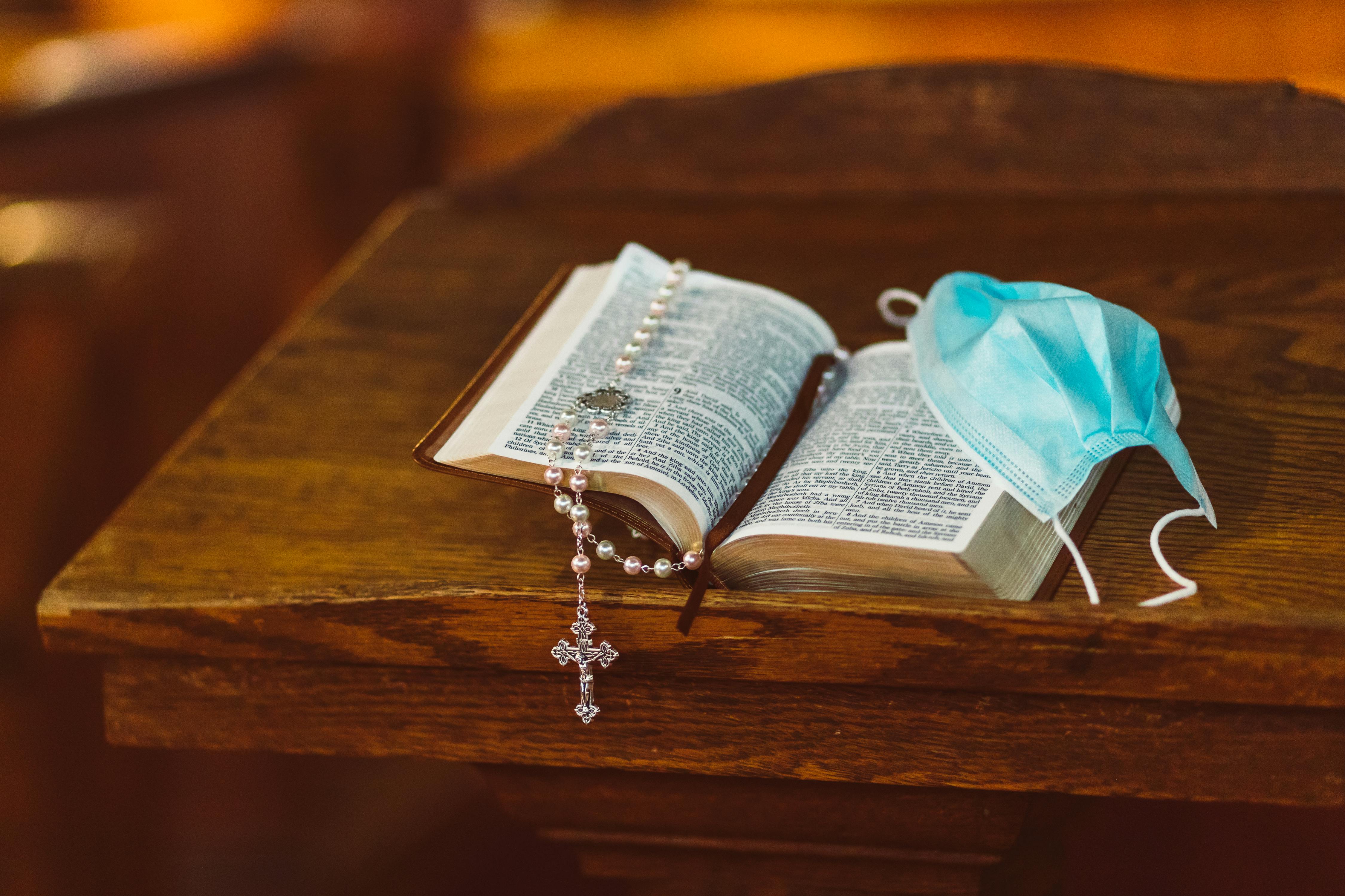 An open Bible with a rosary and face mask, symbolizing faith and current times.