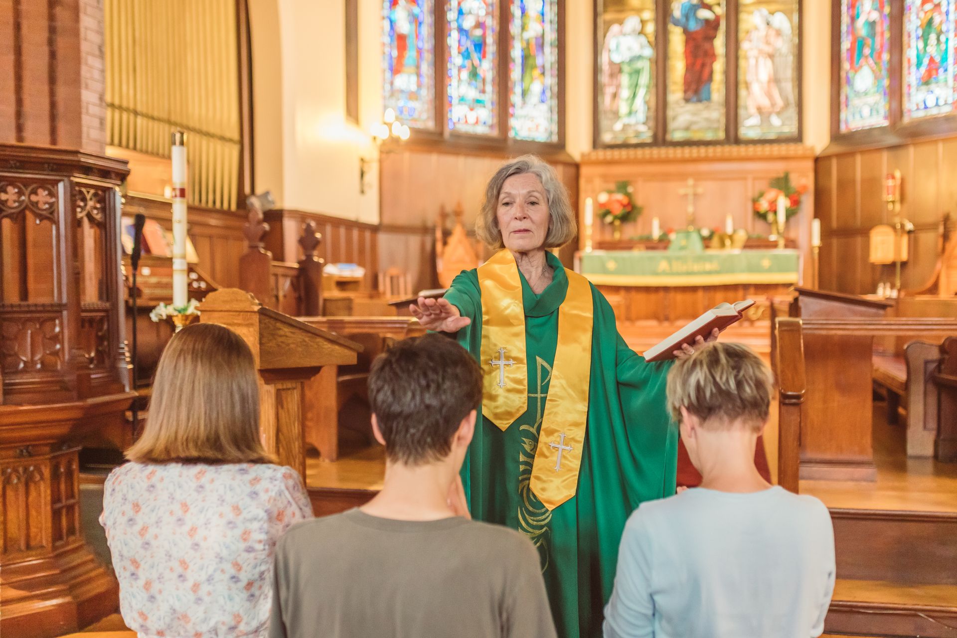 An elderly priest in a green cassock leading a church service with parishioners praying.