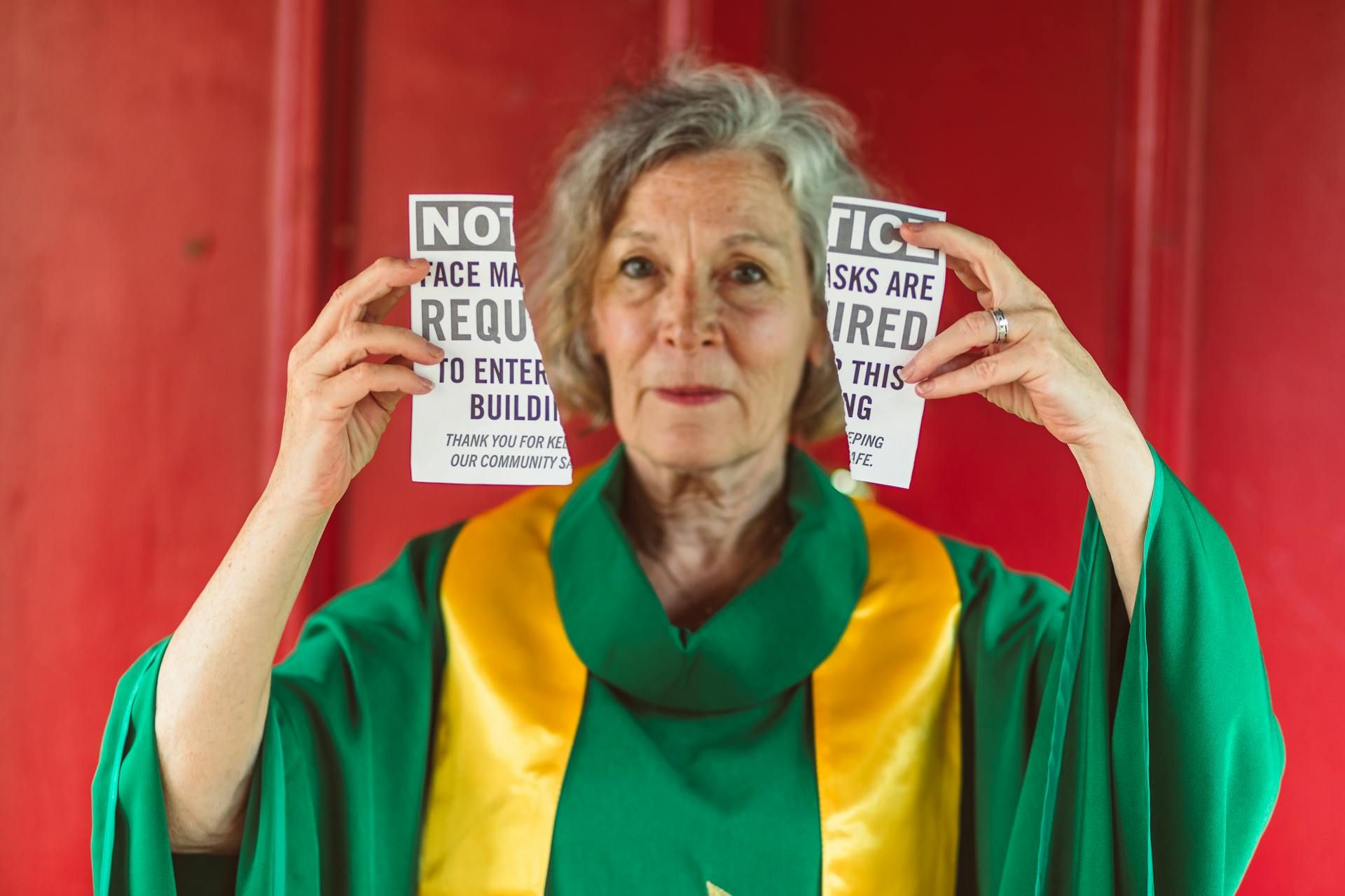 Senior woman in colorful robe holding mask requirement notices against a red background.