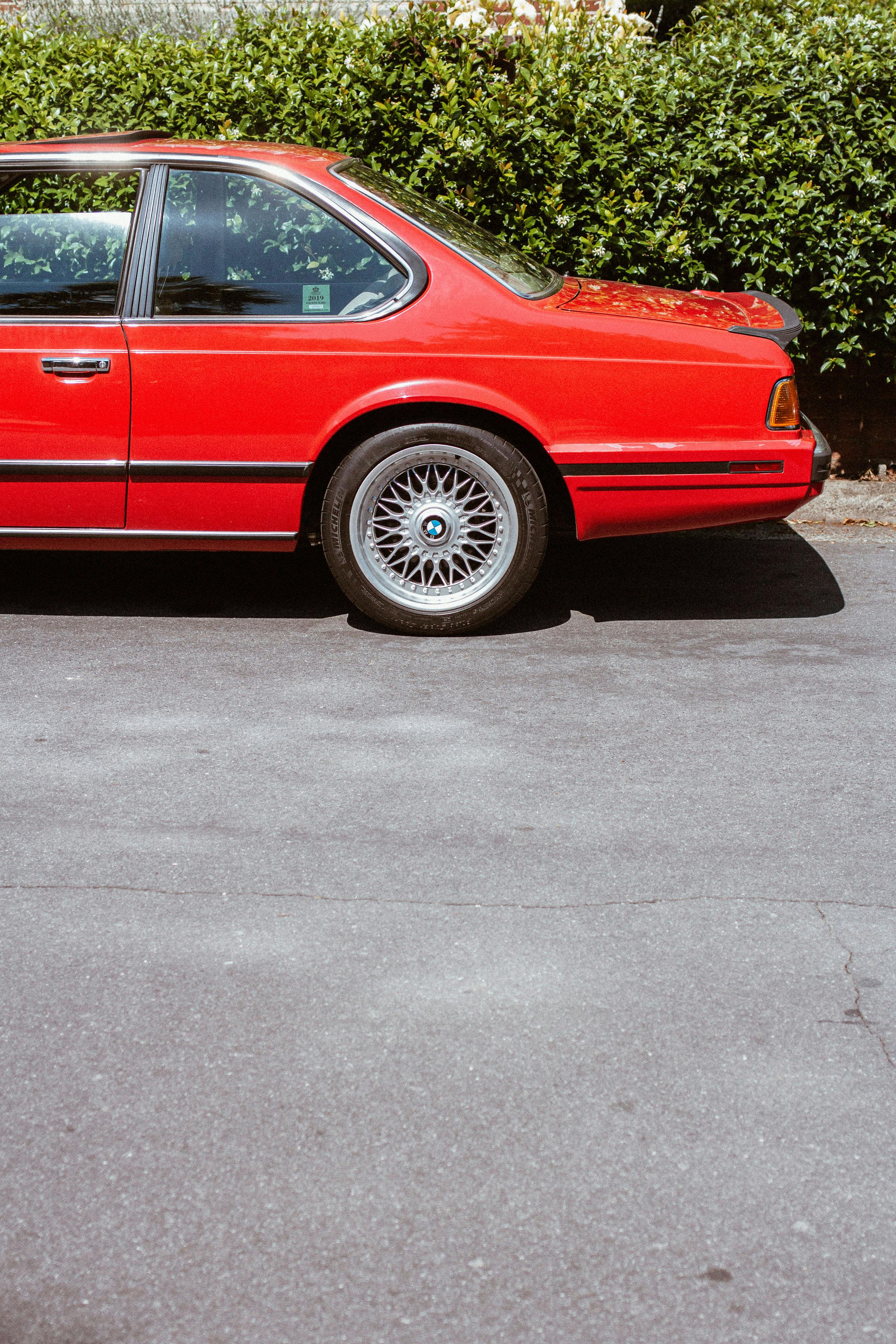 Vintage red car with wire-spoke wheels parked beside green hedges on an asphalt road.
