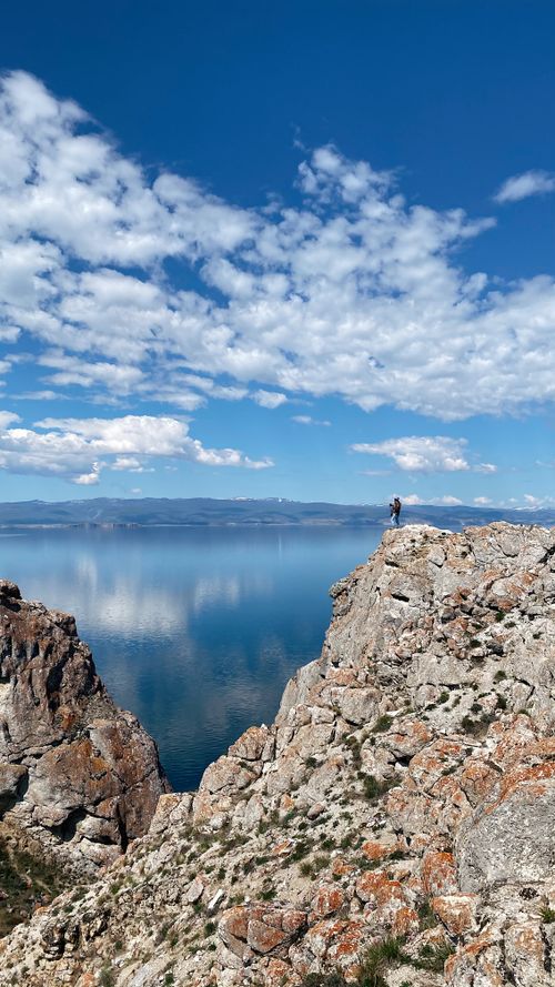 Free Panoramic view of a person standing on cliffs overlooking Lake Baikal under a vibrant blue sky. Stock Photo