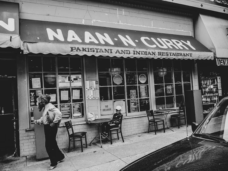 Grayscale Photo Of Man Walking Outside Naan N Curry Restaurant