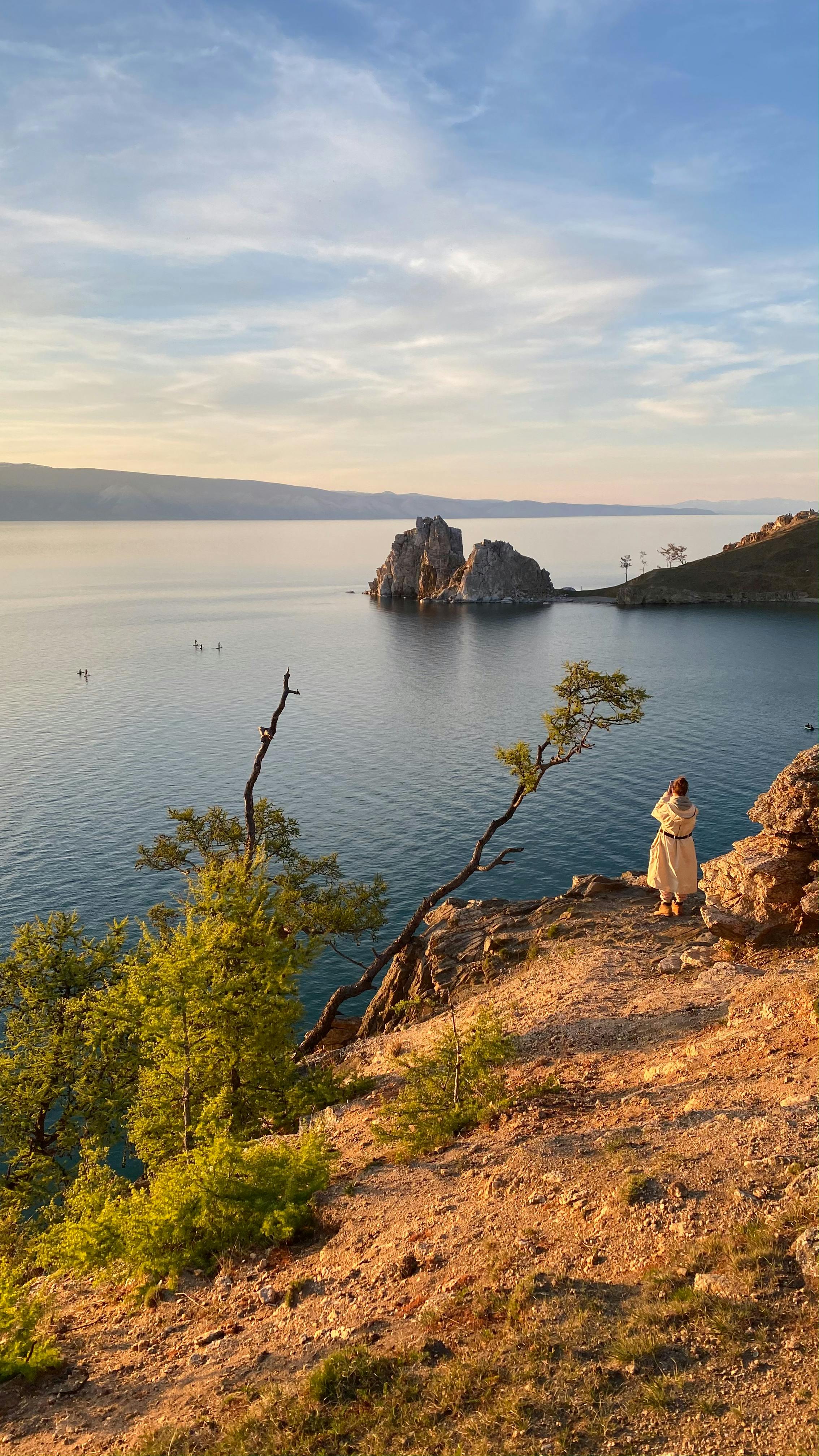Lone traveler gazes over Lake Baikal at sunset, surrounded by scenic cliffs and calm waters.