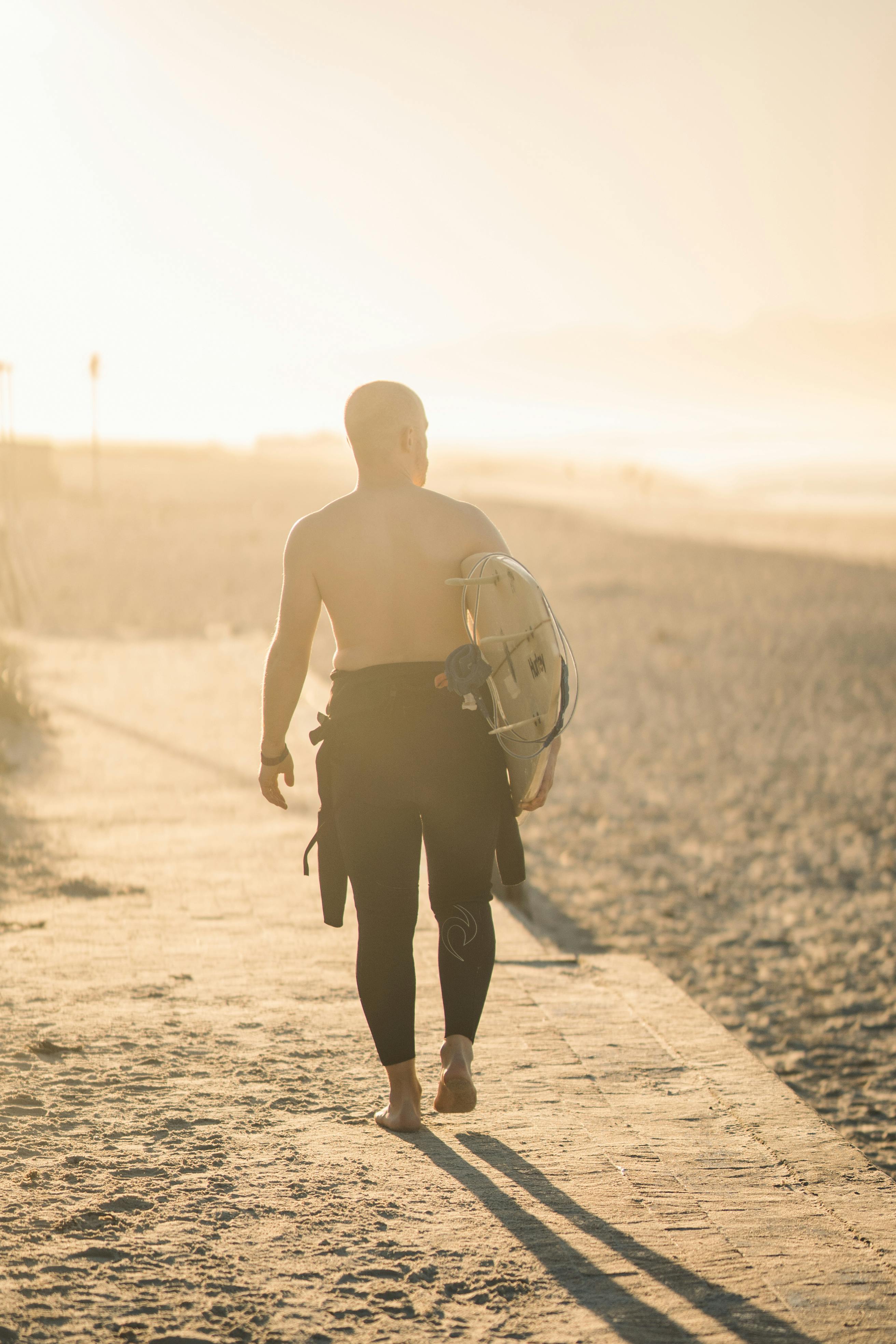 A shirtless surfer carrying a surfboard walks along a sandy beach path under the golden sunset light.
