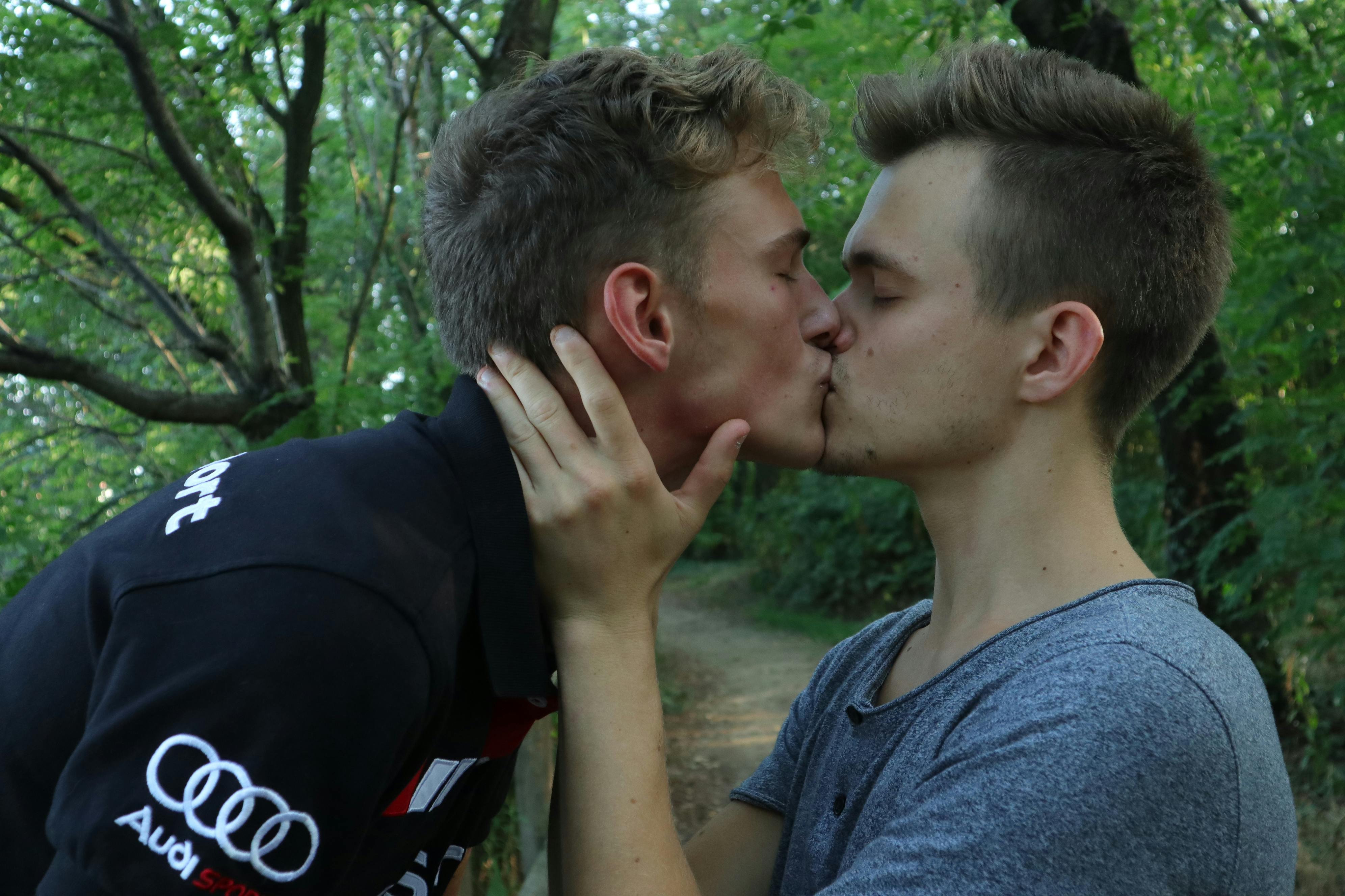 Two young men share a tender kiss in a forest setting, symbolizing love and affection.