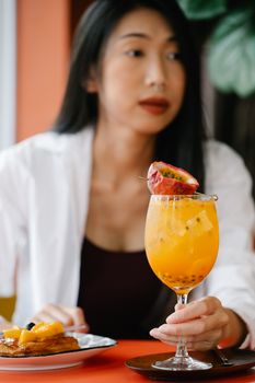 Asian woman savoring a tropical fruit drink in a chic restaurant setting.