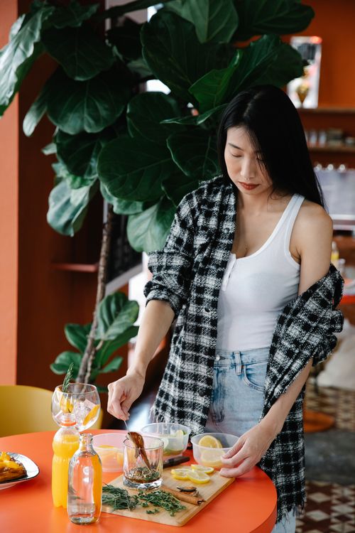 Free Asian woman in plaid shirt and white tank top preparing a healthy lemon herb drink indoors. Stock Photo