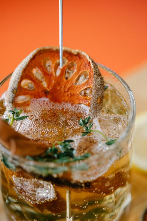 Free Close-up of a refreshing bael fruit cocktail garnished with thyme and ice cubes, in a glass against a vibrant orange backdrop. Stock Photo