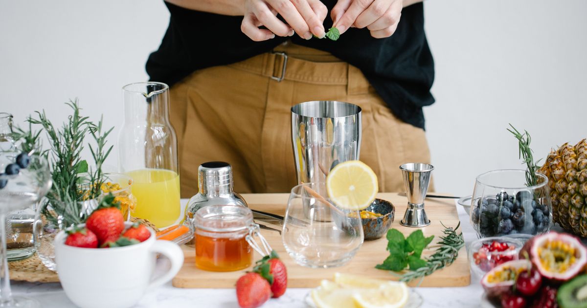 A home cocktail table with a steel shaker, jigger, honey, lemons and fresh fruit, someone adding mint