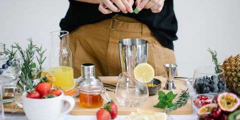A home cocktail table with a steel shaker, jigger, honey, lemons and fresh fruit, someone adding mint