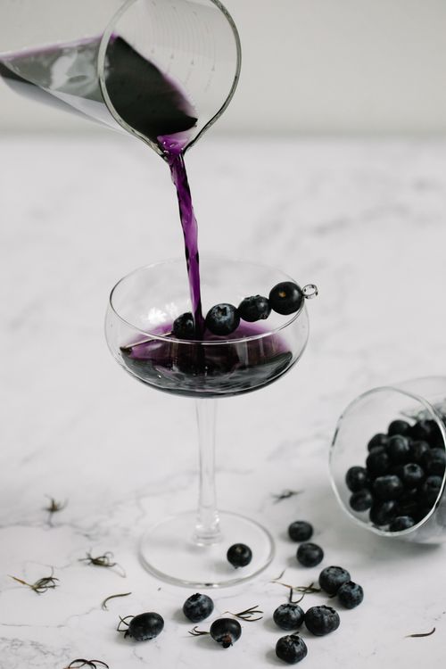 Free A vibrant blueberry cocktail being poured into a glass, surrounded by fresh blueberries on a marble surface. Stock Photo