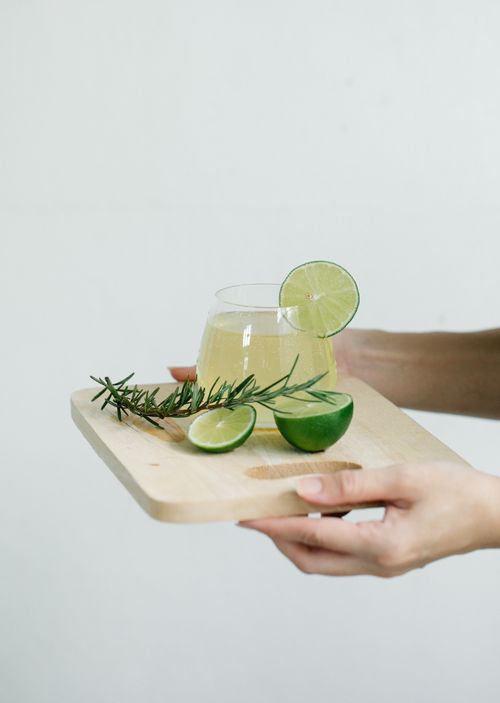 Free Glass of lime juice with rosemary and sliced lime on a wooden board, held by hands. Stock Photo