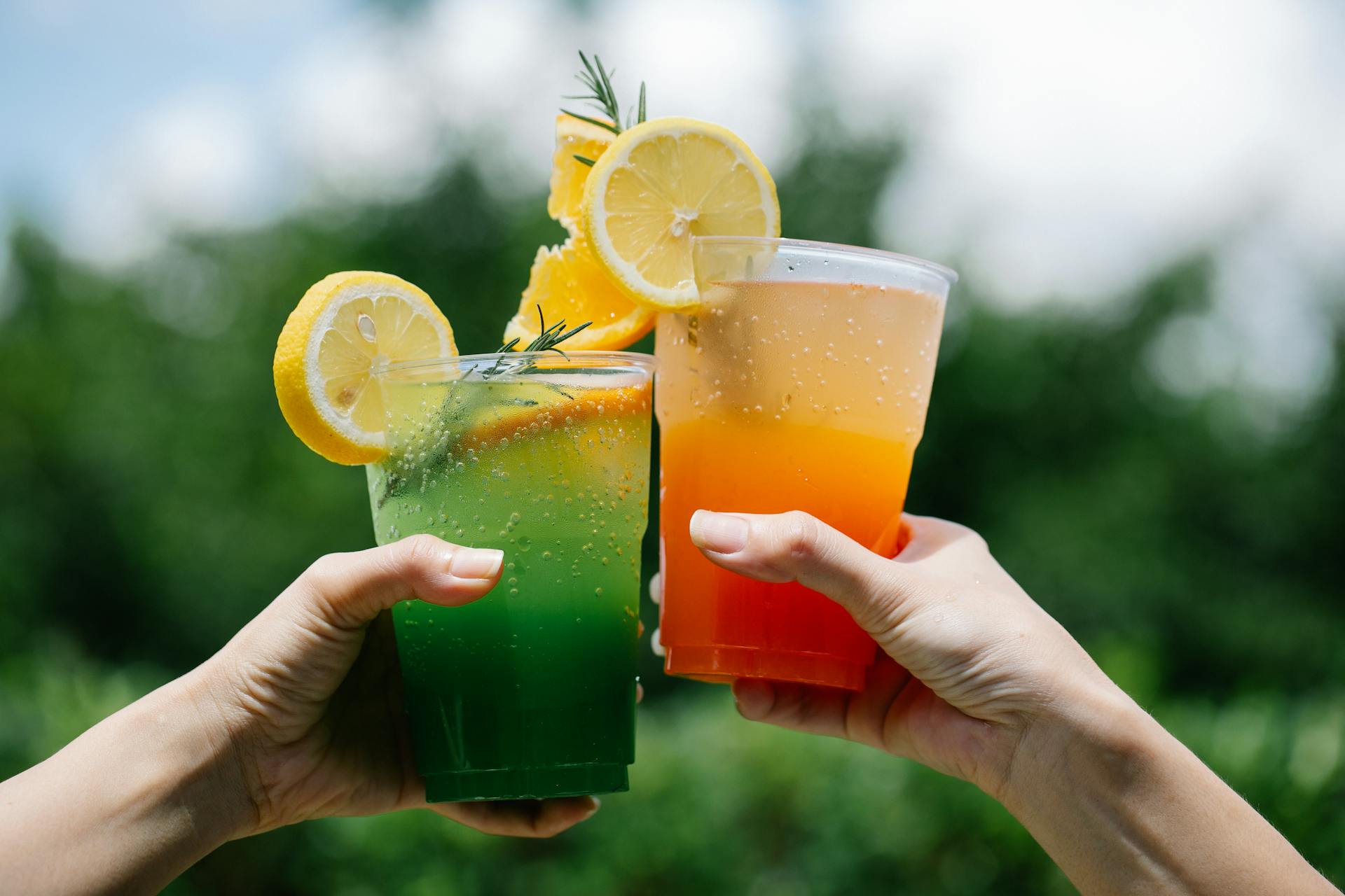 Two hands holding colorful citrus lemonades in a vibrant summer outdoor setting.