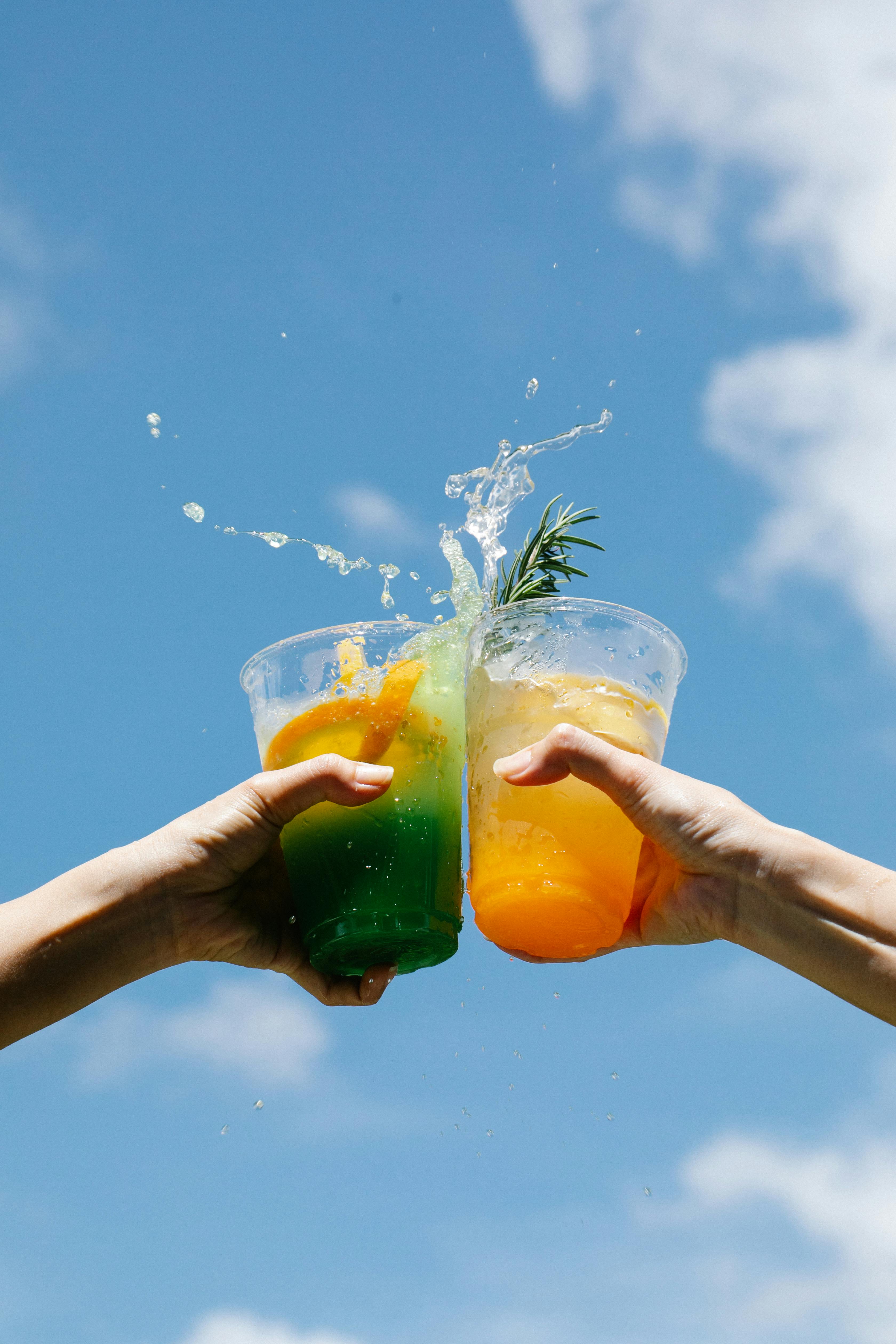 两杯五颜六色的鸡尾酒在明亮的蓝天下叮当作响，时尚地庆祝夏天