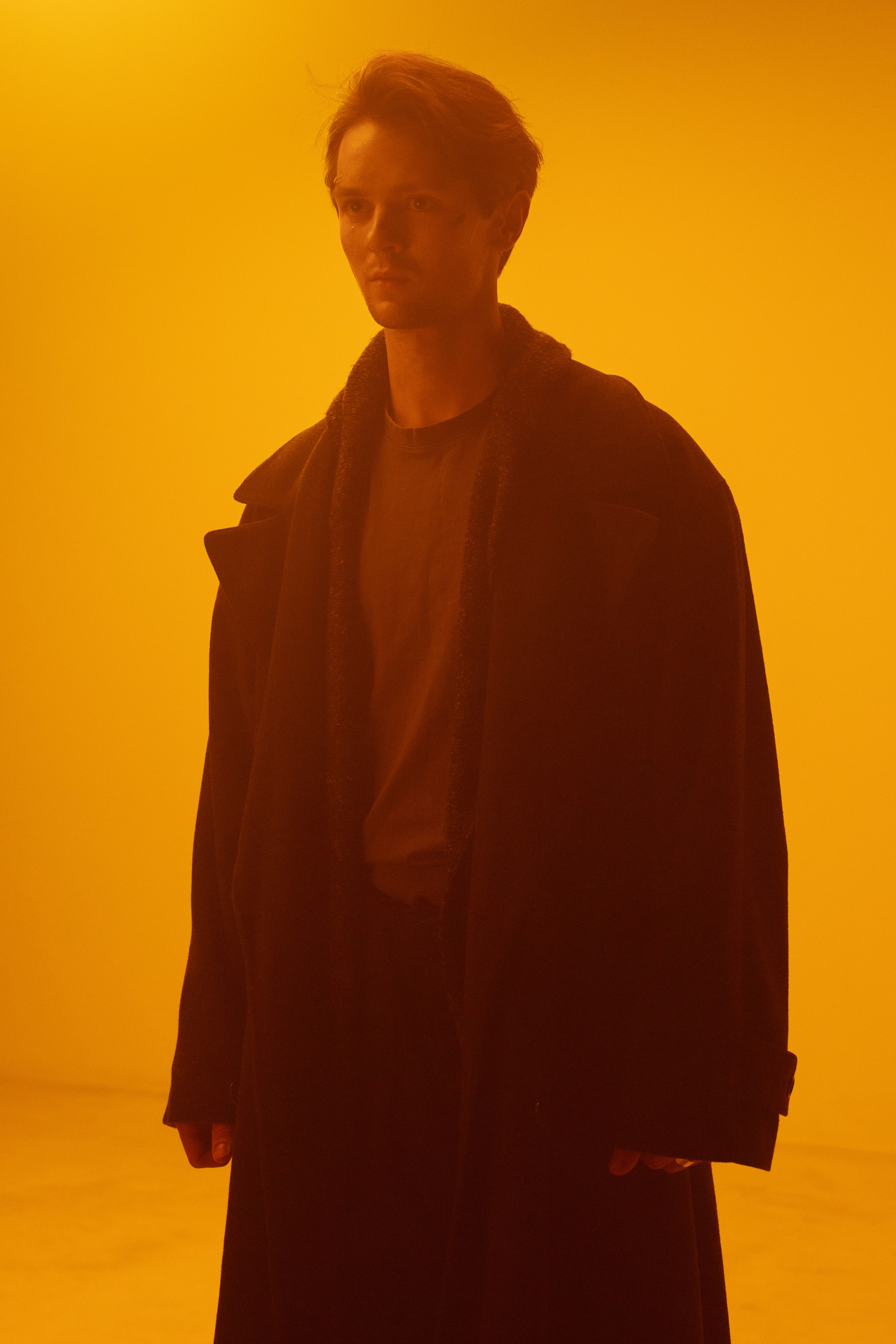 A man stands in an empty studio wearing a long coat, bathed in dramatic lighting.