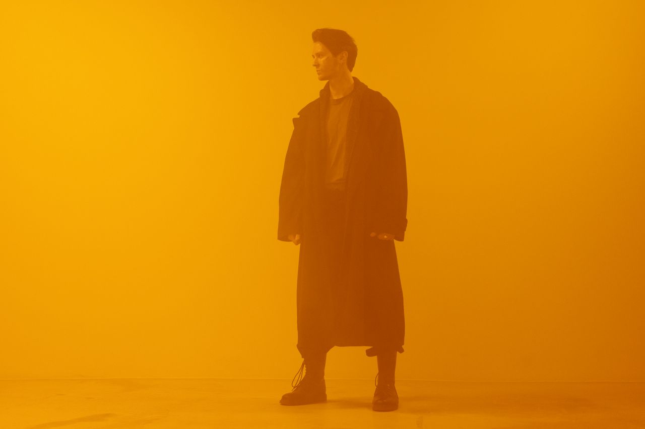 man in black coat standing against an orange background embodying a cy