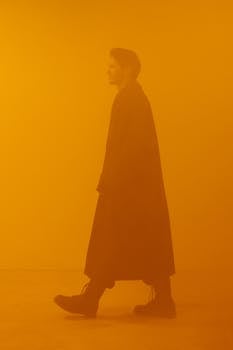 A man dressed in a black coat walks through a conceptual dystopian scene with an orange background.