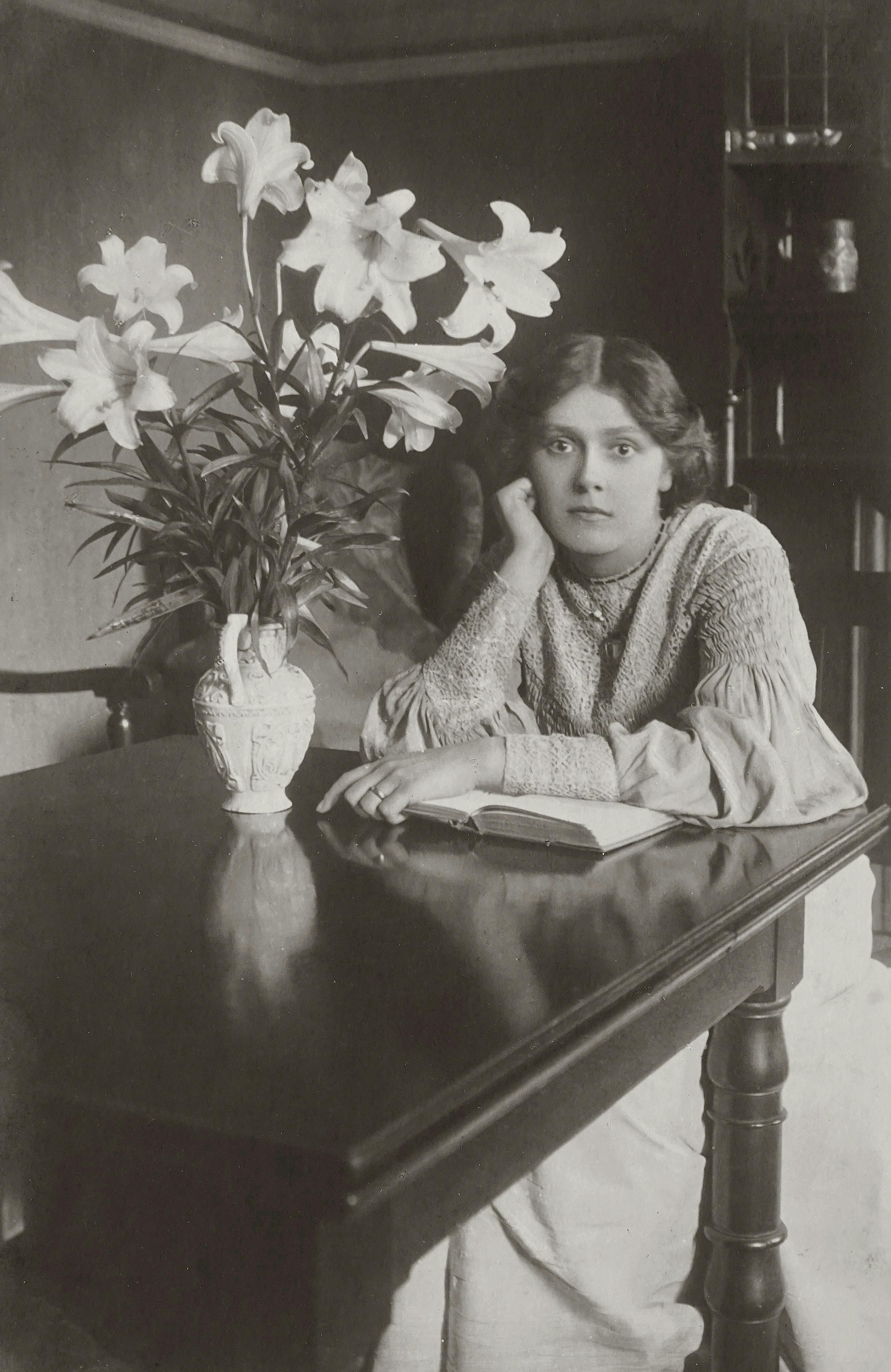 Black and white vintage portrait of a woman with a book and flowers in a vase on a table.