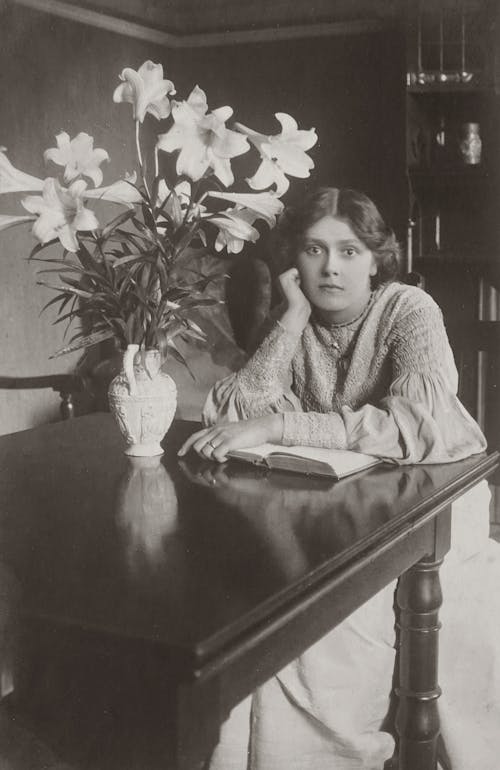 Woman With A Book Sitting Beside A Table With Flower Vase