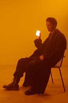 A man in a coat sits on a chair in a dramatic orange-lit studio, emphasizing a dystopian theme.