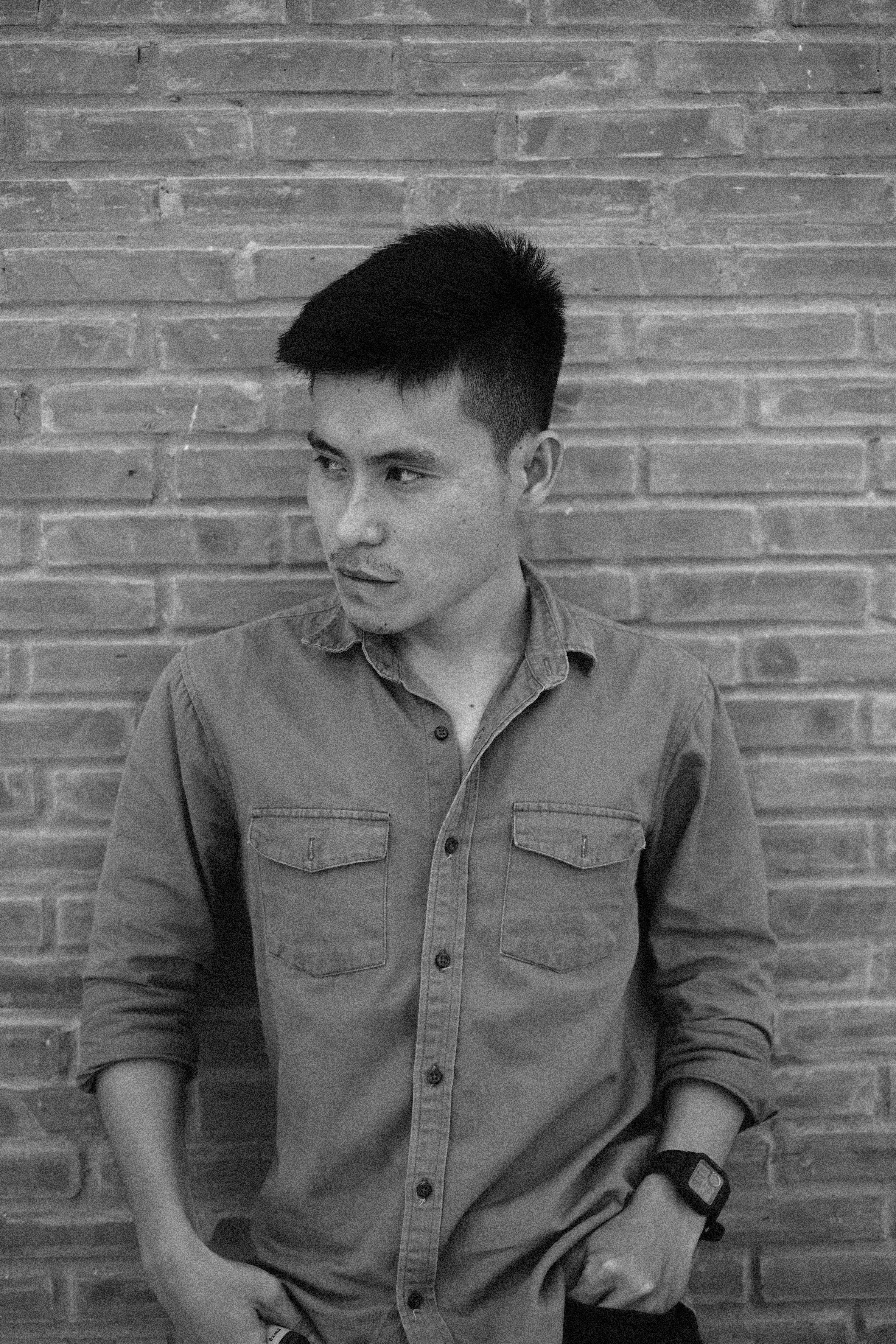Black and white portrait of a stylish young Asian man posing against a brick wall.
