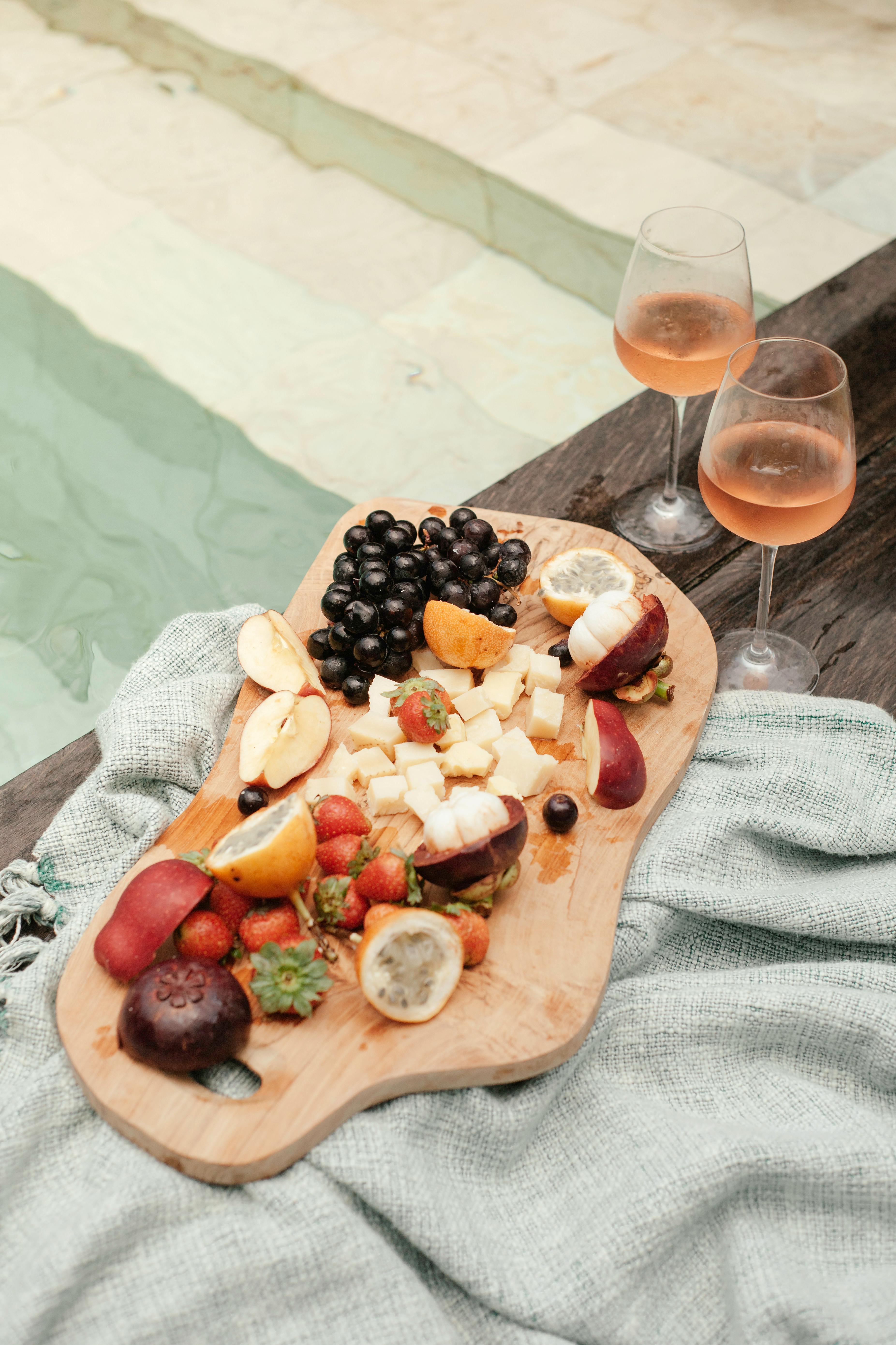 variety of fruit on a cutting board and wine in glasses on the edge of a pool