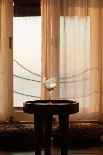 A serene composition featuring a solitary glass on a wooden table by a bright curtain-covered window.