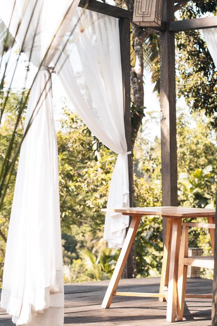 Serene outdoor patio with white curtains and wooden table, nestled among green trees. Ideal for a relaxing escape.