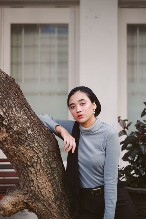 Free A stylish woman in a gray turtleneck leans against a tree trunk in an urban setting, exuding elegance. Stock Photo