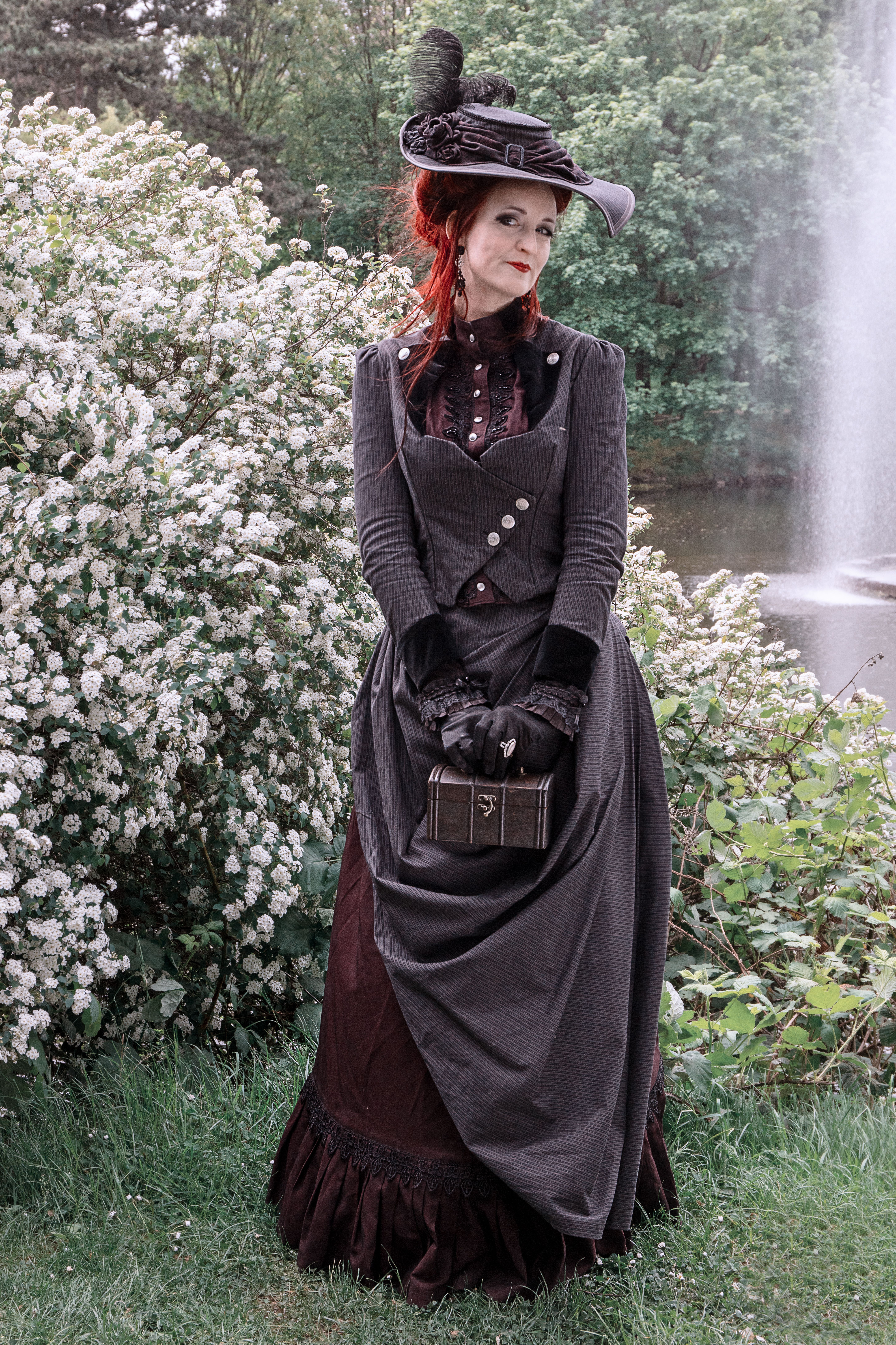 Elegant woman in Victorian attire posing beside lush flowers with a fountain backdrop.